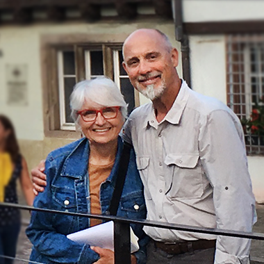 Sherry & John Stilin standing behind a black fence in front of a historic building. The streets are cobbled, the building is white stucko and wood beams. John has his arm around Sherry lovingly, she leans into him and they both smile happily at the camera. She's wearing a tan shirt wiht a denim jacket, her hair is white and cut short around her ears with swooping bangs, and red glasses. He is graying with a white goatee and a pale grey button up outdoors shirt and dark grey pants.