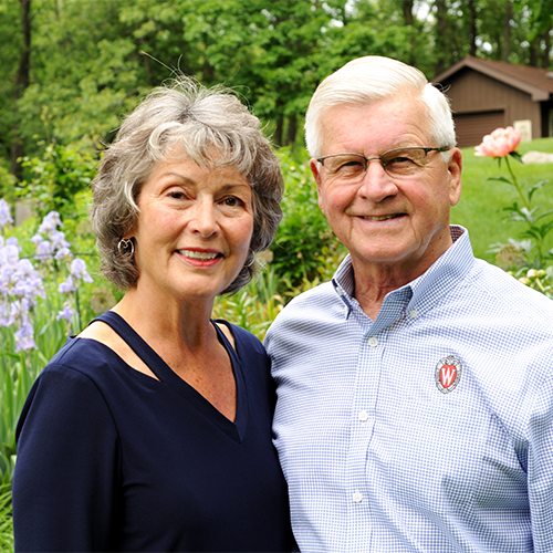 Pat & Al Wilkening standing close together, hugging sideways, outside smiling at the camera.